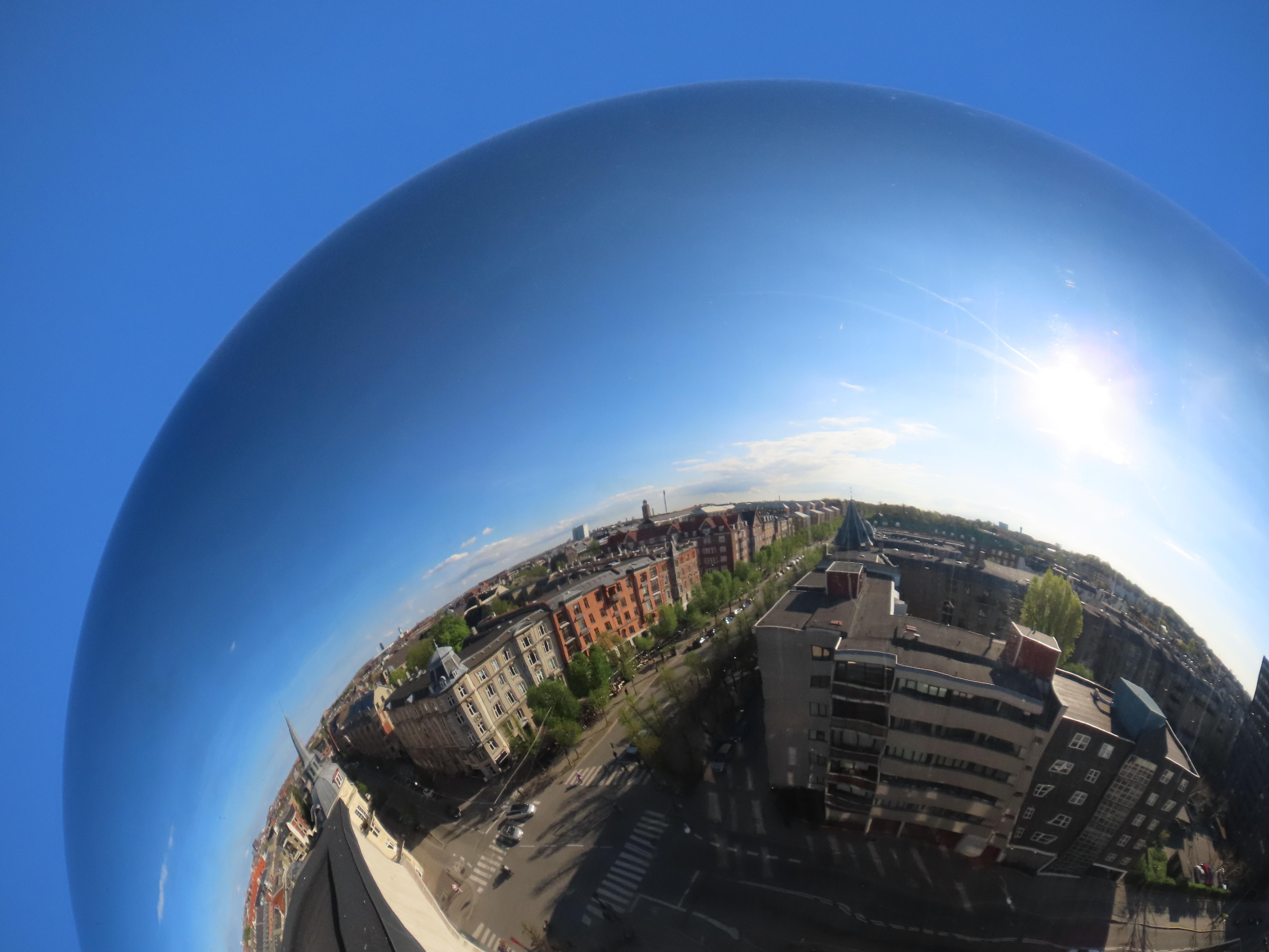 Copenhagen (well, Frederiksberg actually), seen reflected off the surface of a mirrored sphere. The horizon is vertical on the left, then bends to horizonal, then dips about 45 degrees down on the right. The sky is clear bright blue, with just some clouds way in the distance (in the reflection). Behind the sphere, just more clear blue sky.