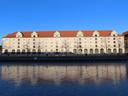 A winter's day: the sky is clear and bright blue. Below that, an old, long warehouse building (mid 1800s?), with a red tiled roof and light beige walls. Then a gravelly courtyard area, in shade. Then the ice covering the harbour, unevenly cracked by the freezing and thawing, and reflecting the colours of the sky and the building.