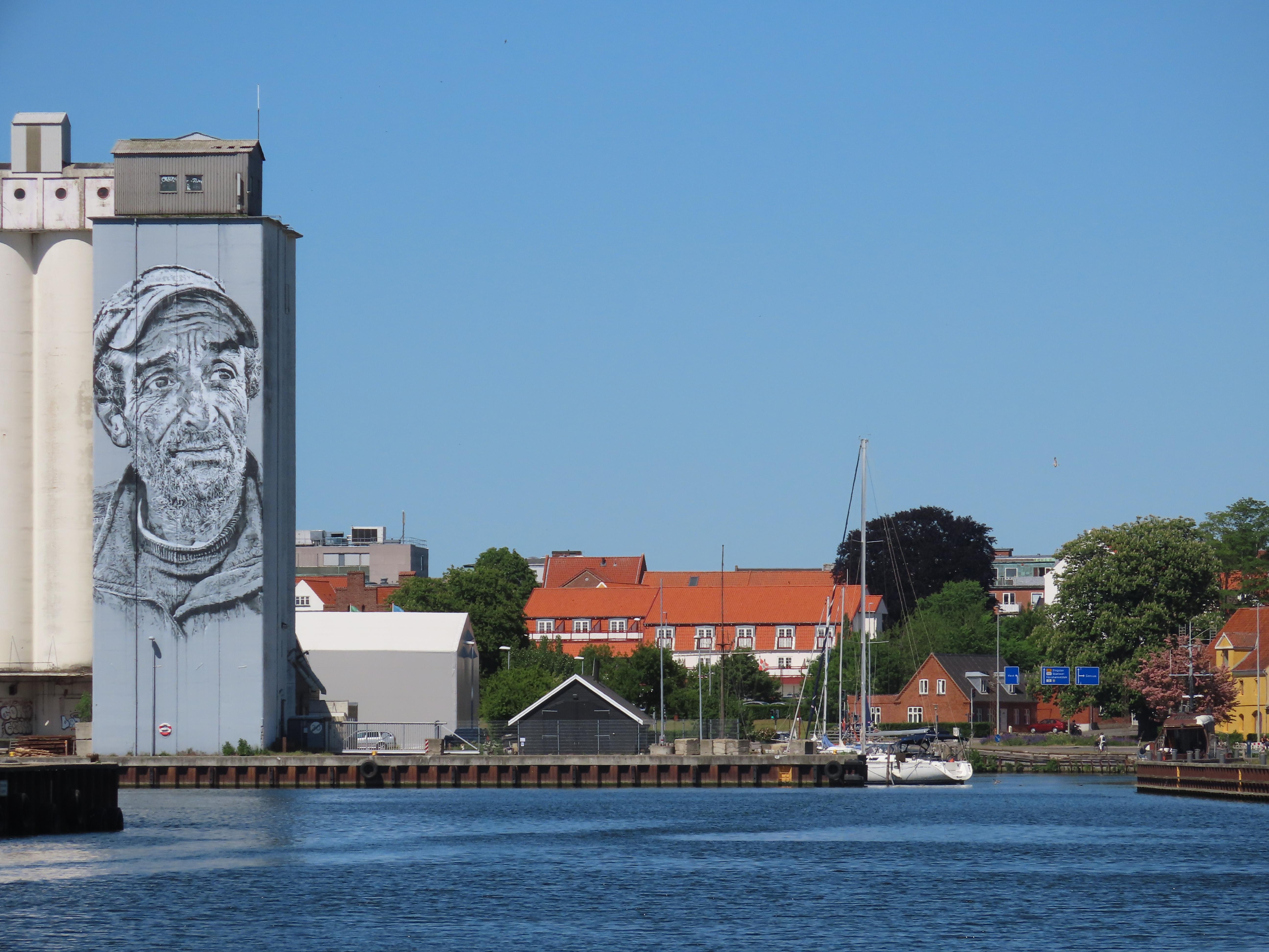 On a lovely sunny day, a small town harbour. Forming the left frame of the picture is a tall metal building (grain tower? I'm no expert), which features a large monochrome painted portrait of a bearded man with a cap — maybe a local fisherman.