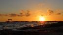 A colourful sunset at the beach. There is a jetty going some 25 meters out to sea, with a bench at the end.