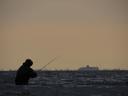 A person in waders (hopefully), standing waist-deep in the sea, holding out a fishing rod. They are completely in silhouette, and facing away from us. In the far distance, on the horizon, but sort of where the fishing rod is pointing, is a ship.