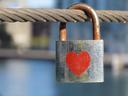 Close-up of a padlock hanging from ~ 8mm stranded wire cable, horizontal. The padlock is significantly rusted and weathered; on the body is a red heart symbol. The background is blurry and mostly blue.