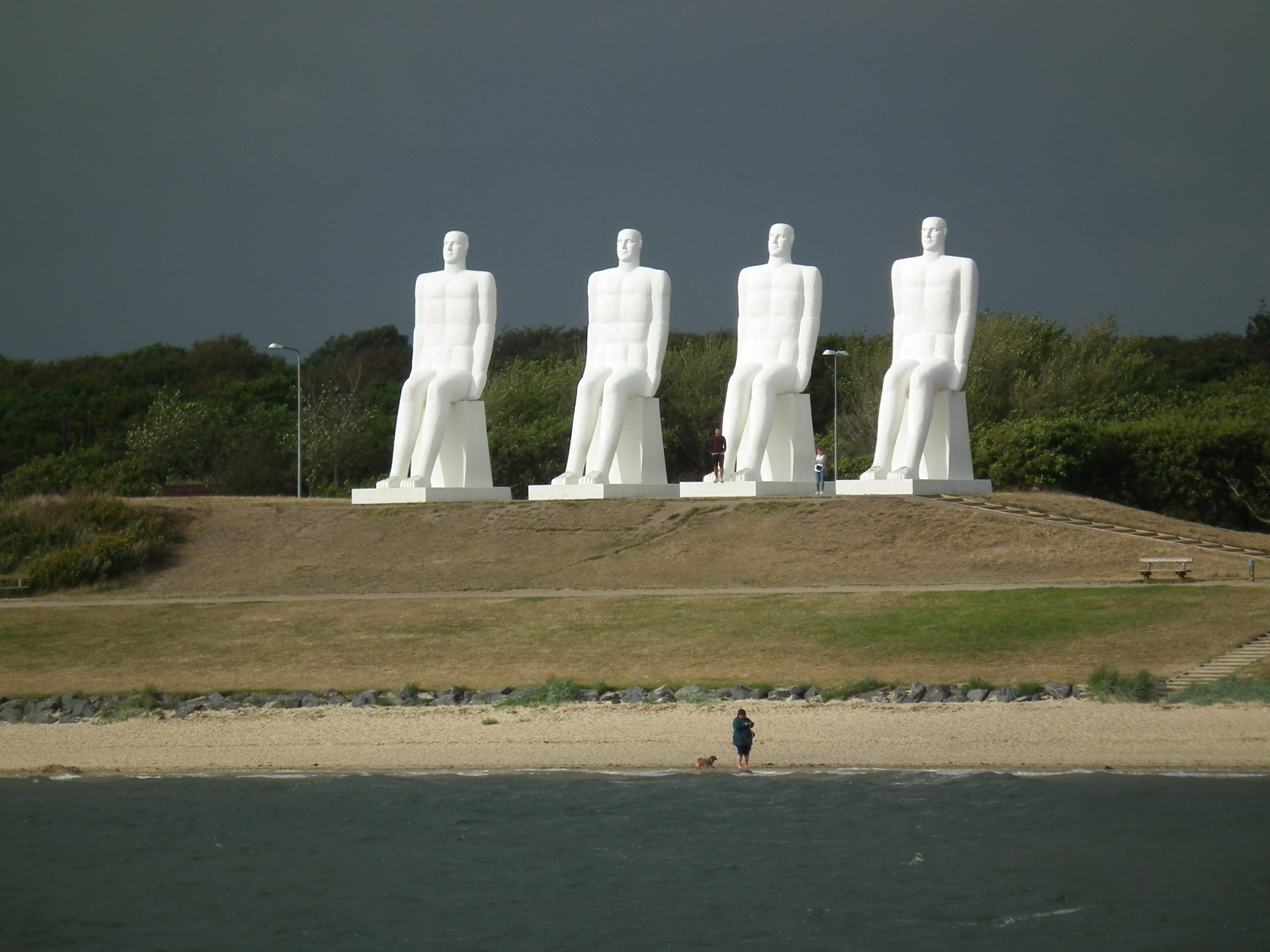 Four large white statues of human figures, sat overlooking the beach, and staring out to sea. Each figure is around 9 meters tall. The all-over brilliant white figures are in stark contrast to the dark grey of a gathering storm cloud behind them.