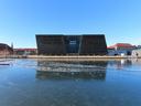 Copenhagen harbour, on a sunny winter's day. There is a completely clear blue sky. We look across the harbour to the city centre: the imposing, black, parallelogram form of the library is directly opposite, flanked by the more traditional red-tiled brick buildings of years gone by. The bottom half of the picture is the frozen-over water, reflecting the reds, black, and blue of the scene. A gash of a path has been cut through the ice, where the harbour bus has sailed through.