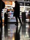 A brightly-lit shop in an airport. The camera is low, so that we can see the dark, shiny floor. In front of the shop is a woman in silhouette, checking her phone. A suitcase is next to her.