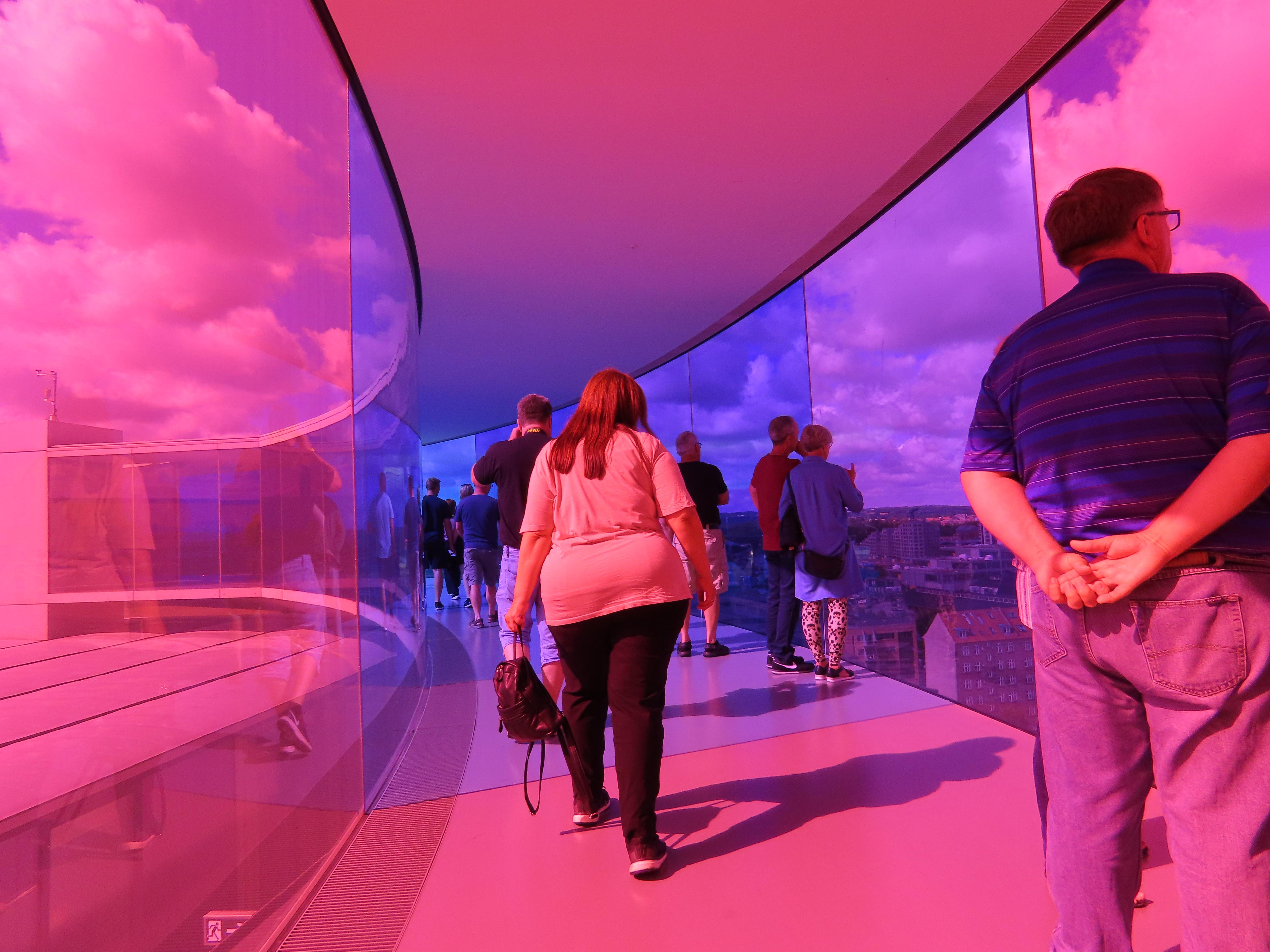 High above the roofs of nearby apartments, a circular glass tunnel, maybe 50m in diameter. There are people inside it (as are we, the viewer). The glass is coloured, with various parts of the circle in shifting shades. This particular part is pink, purple, and blue.