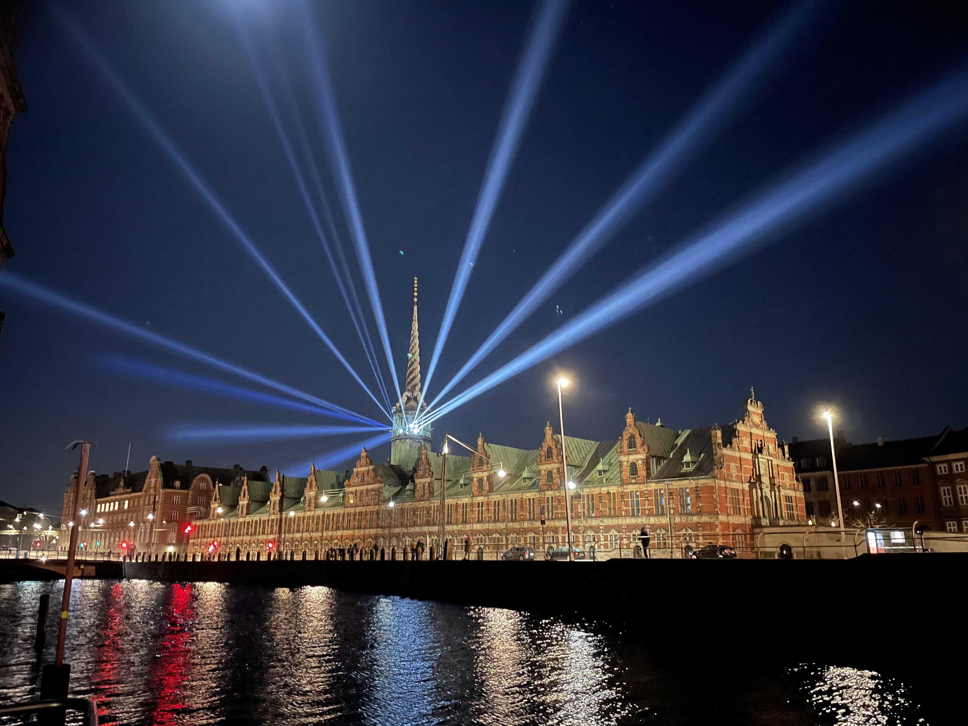 Night time in Copenhagen city centre. The renaissance building 'Børsen', the old stock exchange building, is lit up with rays of blue-white light shooting out from its tower in all directions.